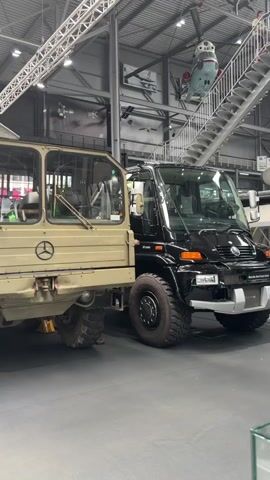The video displays two Mercedes-Benz Unimogs parked indoors, likely in a museum or exhibition hall. The Unimog on the left is a military-style vehicle painted in a tan, desert camouflage color. It features a dual-cab design with visible windows, windshield wipers, and the Mercedes-Benz logo prominently displayed on the front. The tires are large and rugged, typical of off-road vehicles. The Unimog on the right is a more modern, black variant, also with a dual-cab and large tires. Its front grille and headlights are visible, and it also displays the Mercedes-Benz emblem. The background shows a large industrial-style ceiling with exposed beams and lighting fixtures. Suspended from the ceiling are various objects, including what appears to be a helicopter on the right side of the frame and other aircraft components or models. Metal stairs are visible on the right, leading up to a higher level. The floor appears to be a smooth, dark surface. The overall environment suggests an indoor exhibition dedicated to vehicles, possibly with a focus on military or heavy-duty equipment, and potentially aviation. There are no people clearly visible in the frame, and no direct interactions are depicted. The camera pans slightly, providing a static view of the two vehicles.