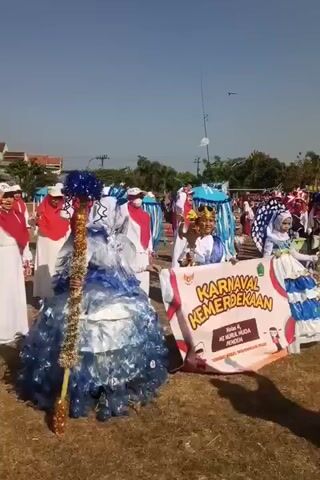 A group of children and adults are participating in an outdoor parade. A young person in a large blue and white costume with a tall staff decorated with blue pom-poms is in the foreground. Beside them, a banner reads "KARNAVAL KEMERDEKAAN" (Independence Carnival) and mentions "Kelas 4 / MI NURUL HUDA PENDEM". Several other participants are visible behind them, some wearing white and red attire with headscarves, others in blue and white costumes. Some individuals are holding umbrellas or parasols. The ground is covered with grass and dry leaves, suggesting an outdoor event on a field. In the background, there are trees and buildings. The sky is clear and blue. A kite is visible flying in the sky. The overall scene depicts a festive community event.