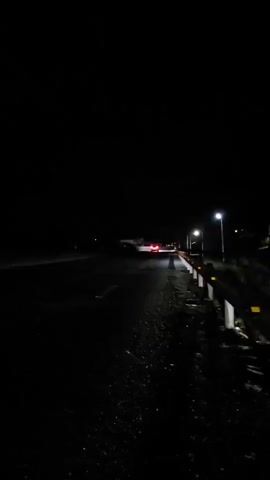 A road at night is seen. The road is dark and wet. A car drives by, followed by a truck and bus. The bus has blue lights. The scene is shot from the side of the road. The road is bordered by a metal guardrail and streetlights.