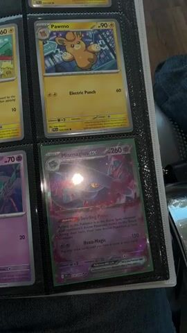 The video shows a close-up view of a black binder filled with Pokemon cards. The binder is open, revealing multiple pages with cards displayed in clear plastic sleeves. The lighting in the room creates reflections on the cards, obscuring some details. 

In the foreground, the bottom right page displays a Mismagius EX card with holographic effects. Above it, a Pawmi card is visible. The page to the left features a Pawmi card, and above that, a Yamper card. Further to the left, a different Pokemon card is partially visible, along with another card below it. 

The top row of the binder shows cards with light blue backgrounds, featuring Pokemon like Piplup and Prinplup. Another Pawmi card is also visible in the top row. The bottom row includes a Pawmot card and another Misdreavus card. The background is dark, with the binder itself being black. The video is shaky and unfocused in places.