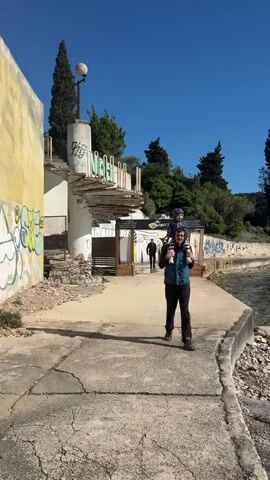 The video depicts a man carrying a young child on his shoulders while walking along a cracked concrete path near the water's edge on a sunny day. Behind them, two people are walking through a wooden gate with a "VIP" sign. To the left, there is a decaying concrete structure with graffiti and a spiral staircase. Trees dot the landscape and a clear blue sky is above.