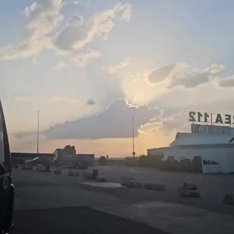 The video opens with a close-up of the side of a dark-colored truck cab, then pans to the right revealing a wide shot of a parking lot at sunset. In the distance, a large cloud partially obscures the setting sun, creating dramatic sunbeams. A white building with a sign that appears to read "AREAS" is visible on the right side of the frame. Several trucks and concrete barriers are scattered throughout the parking lot. The sky is filled with scattered clouds, and the overall lighting suggests late afternoon or early evening.