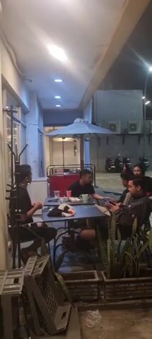 A group of six people are gathered at an outdoor seating area of what appears to be a cafe or restaurant at night. They are seated around two tables. In the foreground, to the left, a person wearing a black t-shirt and shorts is seated. To their right, a person in a black t-shirt is seated at the same table. On the second table, further to the right, three people are seated. One person in a dark jacket is looking at their phone. Another person in a dark jacket is also looking at their phone. A woman in a black top with a pink accent is seated behind them. Further to the right, another person in a black jacket is seated. In the background, there are parked motorcycles and an air conditioning unit mounted on a wall. The area is lit by overhead lights. There are plants in planters in the foreground.