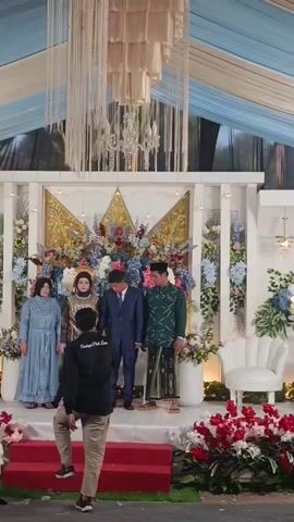 A slightly elevated, wide outdoor shot from a lower angle shows a wedding reception with an elaborate stage setup. In the background, a white stage with gold accents and a large floral arrangement serves as a backdrop. A couple, dressed in formal wedding attire, stands center stage. The bride wears a gold and black gown with a head covering, and the groom is in a blue suit. To their left, an older woman in a light blue dress stands. To the right of the groom, a man in a green patterned shirt and a sarong stands. In the foreground, a man in a black long-sleeved shirt and tan pants is seen from behind, holding a camera and focusing on the couple. The man is on a red elevated platform, which is covered with flower petals. The area around the stage is decorated with various floral arrangements, and a white, ornate chair is visible on the right side of the stage. The overall color scheme of the decorations includes white, blue, gold, and various floral colors. The sky or ceiling above is draped with white and light blue fabric, and a large, intricate chandelier hangs in the center. A man in a dark shirt and pants is standing on a red rug in the foreground, facing the stage.