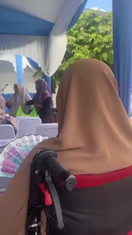 The video shows an indoor event under a blue and white tent. Several people wearing hijabs are seated, facing a stage area. In the foreground, a person in a wheelchair is visible, their back to the camera. The chairs are covered in white fabric, and some have a fan with a floral design on them. In the background, there is a building with a sign that reads "KLINIK DE" followed by other unreadable text. People are interacting and seemingly listening to a speaker or performance. The overall atmosphere appears to be a formal gathering or ceremony.