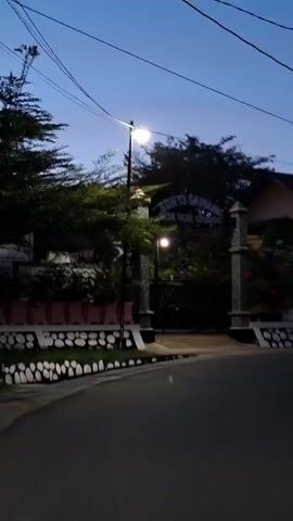 A shot of an outdoor scene, possibly in a rural or suburban area, during dusk or early dawn. A road is visible in the foreground, curving to the right. On the left side of the road, there is a low wall decorated with white circular patterns, followed by a line of trees. A street lamp is illuminated, casting a warm glow on the surroundings. In the background, an archway is adorned with pillars on either side. Above the archway, a sign with text is visible, though it is partially obscured by foliage. More trees and lush greenery surround the archway. Beyond the greenery, a building with a reddish-brown roof and a light-colored exterior is partially visible. The sky above is a deep blue, suggesting the twilight hour. The overall impression is one of a quiet, residential or institutional entrance at the transition between day and night.
