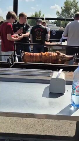 The video shows an outdoor gathering where whole roasted pigs are being prepared and served. In the foreground, a pig is rotating on a spit, slowly cooking. On a wooden surface next to it, two large pieces of roasted pig are visible. One is a whole pig's head, blackened and charred, while the other is a pig's leg, golden brown and glistening. Several people are present, some standing around and observing the cooking, others interacting with the food. One man holds a phone, another is looking at the pigs, and someone is being served food from a tray. In the background, there are grills and more people. The environment appears to be a park or an open-air event.
