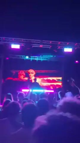 The video shows a nighttime concert with a DJ performing on stage, illuminated by bright, color-changing stage lights and a large video screen.  A crowd of people stands below the stage, with many raising their arms.  Smoke is released, adding to the visual effects. The overall atmosphere is energetic and lively.