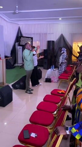 A medium shot shows a gathering in an indoor space. Numerous people, mostly adults of varying ages, are standing among rows of red chairs. Many have their heads bowed or hands raised, appearing to be in prayer or worship. A man is visible on a small stage, possibly leading the service. The room has bright lighting and minimal decor, with a few black draperies in the background. Some speakers are positioned in the back corner. The setting looks like a church or event hall. The attendees are dressed in diverse attire, ranging from casual to formal. Some individuals are embracing or comforting one another. The overall atmosphere suggests a spiritual or emotional event.