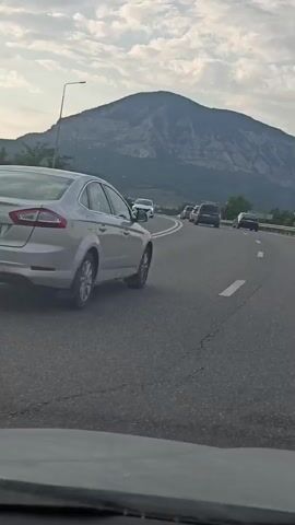 A car is driving on a highway. The car is a light-colored sedan. The highway has multiple lanes and is surrounded by a metal guardrail on the right side. There are other cars on the highway. In the background, there is a large, rocky mountain with a cloudy sky. The sun appears to be setting or rising, casting a soft light. The car's license plate is visible and reads R202XX77. The video appears to be filmed from another vehicle.