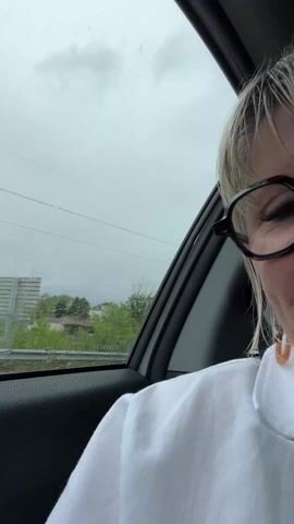 From inside a moving car on a cloudy, rainy day, a woman wearing glasses and a white collared shirt looks mournfully at the camera, and briefly puffs out her lower lip. Rain streaks across the passenger window. The landscape seen through the window includes trees and a few buildings.