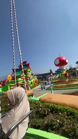A woman wearing a light beige hijab, sunglasses, and a maroon top is seated on a swing ride. The swing is suspended by multiple metal chains from a colorful, striped canopy. The ride is stationary. In the background, there is a vibrant theme park with buildings designed in the style of cartoon characters, including a large red mushroom and multi-colored houses with pointed roofs. The ground is covered in green artificial turf with patches of flowers. Other people are visible on adjacent swings and walking around the park. A large Ferris wheel is also seen in the distance. The sky is clear and blue. The woman smiles at the camera during a selfie-style shot.