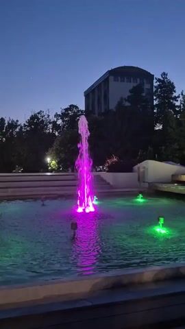 The video shows a fountain with colored lights at dusk. A central fountain sprays water upwards and is illuminated with purple light. Around it, several smaller fountains spray water and are lit with green light. In the background, a large building with many windows and trees are visible against a dark blue sky. The water in the fountain is a bright turquoise color. The lighting of the fountains changes slightly throughout the video, with the purple light sometimes appearing more intense and at other times dimmer. The water jets are mostly static throughout the short clip.