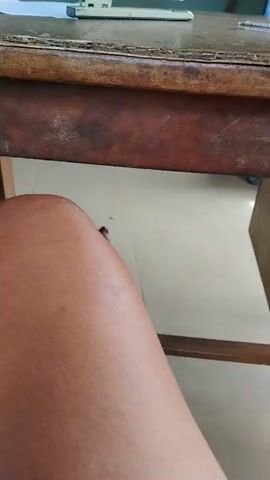 The image captures a low-angle perspective from below a worn wooden desk. In the foreground, the bare legs and feet of a person with brown skin are visible, with red polish on the toenails. The person appears to be seated, with their legs casually positioned under the desk.

The desk itself is made of dark brown wood, showing significant signs of age and use, including scratches, scuffs, and a somewhat faded or uneven finish. A central horizontal beam runs along the underside of the desk. On the desk's surface, several items are placed: a white office stapler, a pen with a green cap, and some white papers.

The floor beneath the desk is tiled with light-colored, possibly off-white or cream-colored, ceramic tiles. A small, dark speck is visible on the floor. To the right, a part of a wooden drawer, consistent with the desk's material, is seen. The scene depicts an indoor setting, likely an office, classroom, or study area, characterized by its functional and somewhat aged furniture. There is no visible text. The general appearance of the desk and tiled floor align with typical interior environments found in regions like Akwa Etiti, Nigeria.
