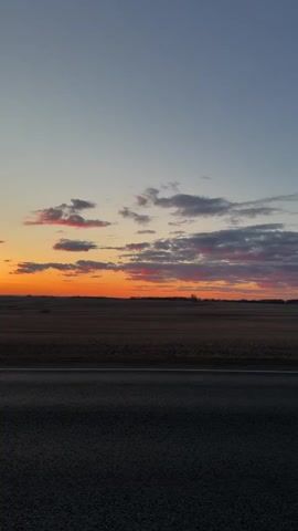 The video presents a static shot of a rural landscape under a colorful sunset. The foreground is dominated by a dark asphalt road, stretching horizontally across the frame.  Beyond the road, a vast, harvested field extends towards the horizon, its surface a mix of brown and tan hues. The most prominent feature is the sunset, displaying a vivid orange band along the horizon, gradually transitioning to pink and gray clouds scattered across the blue sky. The sky occupies the upper portion of the frame, providing a backdrop of calm and cool tones above the fiery sunset.