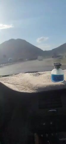 The video is filmed from inside a vehicle looking out through the windshield. The perspective is from the dashboard, which is covered with a light-colored fuzzy mat. A blue-colored car air freshener is visible on the right side of the dashboard. The road ahead is visible, with a guardrail on the left. Several vehicles, including cars and a truck, are on the road in the distance. The landscape consists of large, barren hills or mountains under a bright, sunny sky with some scattered clouds. The sun appears to be low in the sky, casting a hazy light. The overall impression is of a scenic drive on a highway.