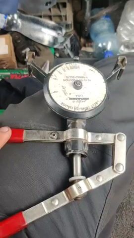 A close-up, top-down shot focuses on a belt tension gauge. The gauge has a circular dial with markings and a needle. Three adjustable arms extend from the top of the gauge, each ending in a clip. A mechanism with red-handled levers is attached to the bottom of the gauge. The gauge and mechanism are resting on a dark-colored, textured surface, likely fabric. In the background, out of focus, are various objects, including plastic bottles, car parts, and a cardboard box. A hand with red-handled levers is visible in the lower left corner, holding and operating the device. The needle on the gauge moves as the levers are manipulated.