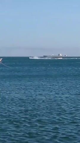 Here is a concise description of the video: 

The video depicts a daytime scene on the ocean. A small fishing boat, painted green and white, is visible in the foreground, gently bobbing on the water. A rope extends from the boat out into the water. In the background, a jetski speeds across the water, creating a wake. Further back, a large cargo ship is visible on the horizon. The sky is clear and blue.