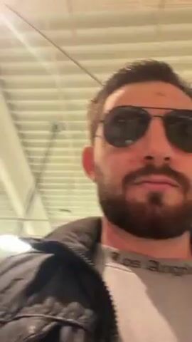 A man with a beard, wearing a black leather jacket and sunglasses, walks through a store. The store has bright overhead lighting and red signs in the background. The man speaks to the camera.