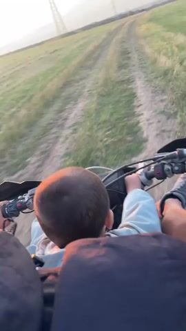 This media file provides a first-person perspective of a motorcycle ride with two individuals. The subjects are two people, one adult and one child, positioned on a dirt bike. The adult is in front, operating the motorcycle, with the child seated behind them. They are riding on a dirt path through a grassy field.

The scene is set in an open, rural landscape, likely in Bulgaria, given the location context of Zheglartsi. The activity is recreational, a motorcycle ride. Notable details include the time of day, which is sunset, indicated by the warm, orange glow in the sky and the low angle of the sun. The weather appears to be clear and pleasant for outdoor activity. There are also power lines visible in the distance, a common feature in rural or semi-rural settings.

The overall emotion conveyed is one of shared enjoyment and adventure, suggested by the perspective and the apparent activity. The child's presence behind the adult hints at a family outing or a bonding experience. There is no visible text within the media file.