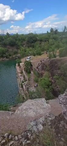 The video shows a quarry with a body of water. There are several people present, some on the edge of the quarry and others near the water. One person is seen sitting on a ledge overlooking the water. A white car is parked in the distance. The environment is rocky with trees and vegetation.