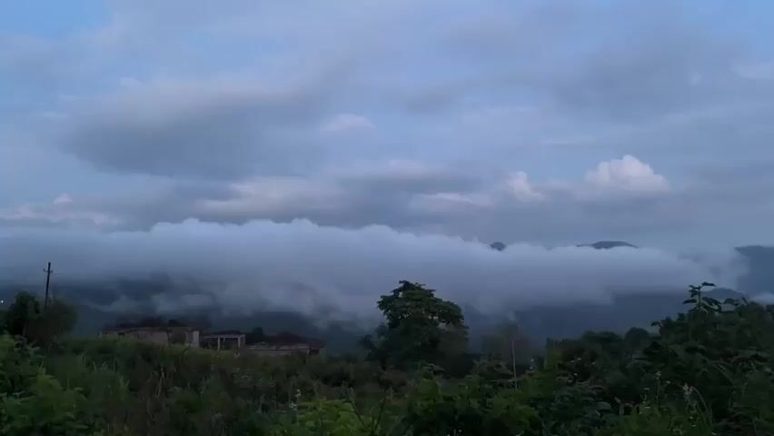 A wide shot captures a misty mountainous landscape under a cloudy sky. In the foreground, lush green vegetation grows, with a few dilapidated structures visible in the middle ground. A dense band of white clouds hangs low, obscuring the middle and distant mountains, creating a sense of depth and mystery. The overall mood is serene and slightly melancholic.