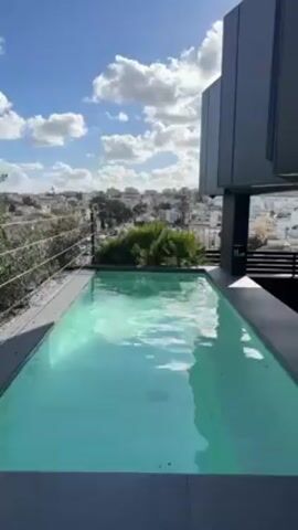 The video shows a rooftop pool with a view of the city. The pool is rectangular and has light blue water. The surrounding area is tiled and has a metal fence around it. In the background, there are buildings and trees. The sky is blue with white clouds. The camera pans from left to right to show more of the city view.