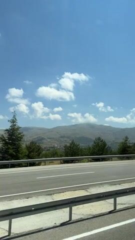 The video shows a view from a vehicle driving on a highway. The road is paved and has white lane markings. A guardrail runs along the side of the road. A traffic sign with a red and white chevron pointing right is visible. Beyond the road, there are green trees and rolling hills with a mountain in the background. The sky is blue with scattered white clouds. A black car drives past in the opposite direction, followed by a white van. Another car, a light-colored sedan, drives past in the same direction as the viewer. A portion of a white car is visible in the foreground, partially obscuring the view.