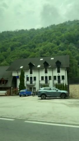 A vehicle is traveling along a road, passing by a large, tree-covered mountain. On the left side of the road, a multi-story white building with a dark roof and visible dormer windows is seen. A dark-colored car is parked in front of it. Next to the white building is a modern, dark-colored building with large windows and a stone wall in front. A large grey dumpster is visible in front of the stone wall. Further down the road, a gas station with a red and black canopy is visible. The gas station has several pumps and a small building with windows. A flag with a logo is hanging from a pole at the gas station. The vehicle continues to move, passing by another building with green trim and diamond-shaped windows, where several cars are parked. The video ends as the vehicle continues down the road.