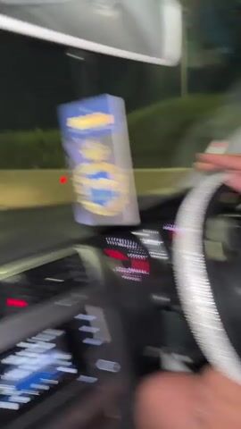 The video shows someone driving a car at night. The driver is holding a cigarette in their hand and has a silver, rhinestone-covered steering wheel cover. There's a car freshener hanging from the rearview mirror. The car is on a freeway, and you can see other cars' lights in the distance.