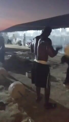 The video captures a dimly lit outdoor scene, possibly at dusk or night, with a group of men engaged in what appears to be physical training. The ground is covered in dirt or sand. In the foreground, a shirtless man with dark skin is visible, wearing dark shorts with a white stripe. He is in motion, possibly lifting or carrying a large, round, pale object. His back is to the camera, and he appears to be sweating. To his right, another shirtless man is partially visible, also in motion. Further back, other individuals are present, though their actions and appearances are less clear due to the low light and blurry quality of the video. Some appear to be observing or participating. There are structures in the background that suggest an open-air facility or an area under construction. The overall atmosphere is one of intense physical activity in a somewhat rudimentary setting.
