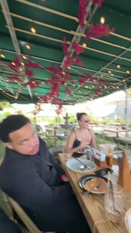 A group of four people are at a table outdoors, under a green awning decorated with pink flowers and string lights. The table is set with plates, cutlery, glasses, and condiments. Food items include pancakes, waffles, and what appears to be eggs. The four individuals consist of one male and three females. The male is seated at the end of the table, dressed in a black jacket, and looks toward the camera. The women are dressed in black tops. One is looking at her phone, another is adjusting her hair, and the third is speaking. The environment suggests a brunch setting at a restaurant with outdoor seating. Additional tables and umbrellas are visible in the background, indicating a public or semi-public space.