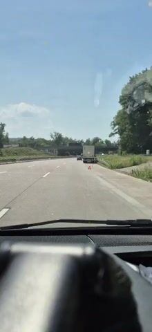 The video shows a view from inside a vehicle, looking through the windshield. The vehicle is on a highway, with multiple lanes visible. To the right, a semi-trailer truck is stopped on the shoulder, with a red triangular warning sign placed behind it. Further ahead, another vehicle is visible in the distance. Trees and vegetation line the right side of the highway, and a bridge or overpass is seen in the distance on the left. The sky is blue with some scattered clouds, suggesting it is daytime. The focus of the camera is on the road ahead, with the dashboard of the vehicle partially visible in the foreground.