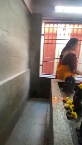 In a dimly lit interior, a woman dressed in a traditional Indian sari, a deep maroon blouse and a vibrant yellow pallu, is engaged in prayer. She stands before a stone altar adorned with small, dark statues, possibly idols of deities, and scattered yellow and red flowers. A tiny flame flickers in a small dish on the altar, suggesting a ritual offering or a lamp. The woman's hair is tied back in a braid, and she appears to be carefully placing an item, possibly a flower, at the base of one of the statues.

The setting appears to be a temple or a shrine, with barred windows revealing a glimpse of the outside world. Through the windows, we see glimpses of red and white striped awnings, suggesting a bustling outdoor market or a brightly decorated area. People can be seen in the background, some in colorful attire, hinting at a lively atmosphere. The interior walls are made of concrete, and the floor is a textured stone. A fluorescent light fixture is visible on the ceiling, providing the primary illumination. The overall ambiance is one of reverence and devotion, juxtaposed with the muted activity visible beyond the windows. The light filtering through the bars creates a pattern of shadows and highlights on the altar and the surrounding area.