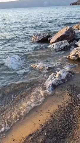 The video shows a shoreline with calm water lapping against rocks and sand. The water is clear and reflects the light, creating ripples. The rocks are of various sizes and colors, with some appearing wet and others dry. The sand is a light brown color, with small pebbles mixed in. In the background, there is a body of water that stretches to the horizon, with a faint line of land visible in the distance. The sky is clear and a light blue, with hints of the sun setting. The scene is peaceful and serene, conveying a sense of natural beauty and tranquility.