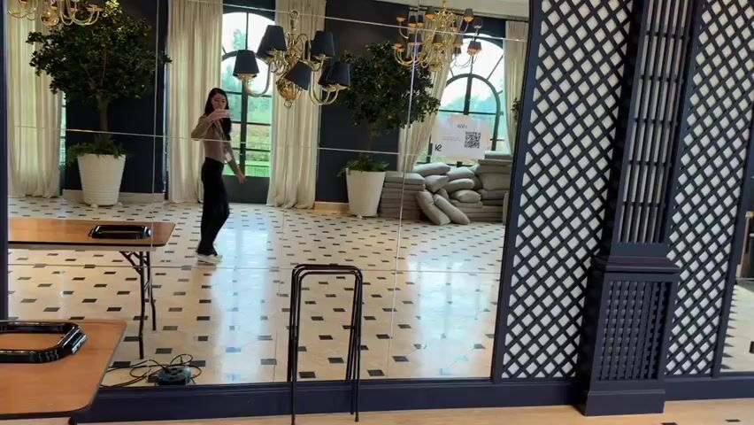 A woman stands in a room with a tiled floor and mirrored walls. She is taking a picture of herself in the mirror with her phone. The room has large windows with white curtains, chandeliers, and potted trees. There is a table to the left of the woman, and a black lattice structure to her right. A sign with a QR code is visible in the mirror behind her.