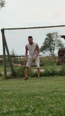 A man is practicing soccer in a grassy field. He is wearing a white sleeveless shirt and white shorts. The man is in the process of kicking a soccer ball. A net is behind him, suggesting he is practicing in a soccer goal. The field is green and lush, with some clover visible. In the background, there is a wooden fence, trees, and a wooden structure. The sky is overcast, suggesting a cloudy day. The setting appears to be outdoors in a rural area. The man's posture indicates he is engaged in a sporting activity. There is no visible text in the media file.