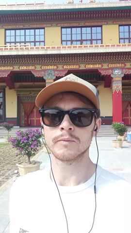 A man in a white shirt, brown baseball cap, and sunglasses is standing outside a large, ornate Tibetan temple. The temple is yellow with red accents and has multiple windows. The man is wearing earphones and appears to be talking to the camera. There are plants in pots on either side of him. The sky is blue and clear.