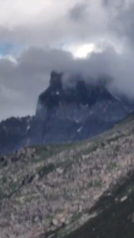 A video shows a mountainous landscape with a jagged peak partially obscured by clouds. The foreground is a rocky, sloped terrain with patches of green vegetation. The sky is overcast with a mix of dark and light clouds, with occasional glimpses of blue sky. The camera pans slowly across the scene, revealing more of the vast mountain range. There are no people or animals visible in the video.