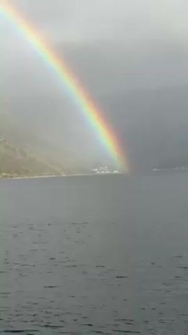 The video shows a double rainbow ending in the water in front of a coastal town. The water is choppy and dark, and the sky is overcast. The rainbow is bright and clear, and the second, less intense rainbow is visible above it. The town is small and white, and it is nestled against the side of a mountain.