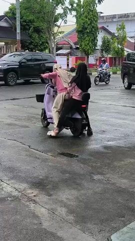 Two young women wearing hijabs are riding on a red electric scooter, one behind the other. The woman in front is wearing a white hijab, a pink jacket, and beige pants. The woman in the back is wearing a black hijab, a pink jacket, and dark pants. They are riding on a wet asphalt road. In the background, there are buildings, trees, and other vehicles, including a black car and another scooter. The scene appears to be in an urban environment.