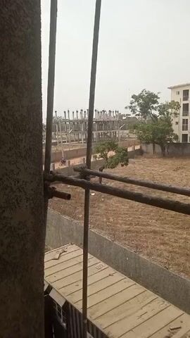 From behind metal scaffolding, the camera pans right across a dry field bordered by a concrete wall. Beyond the field are buildings, including a half-constructed skeleton of a large building and a completed, multi-story apartment building. A water tower is visible in the distance. The sky is overcast.
