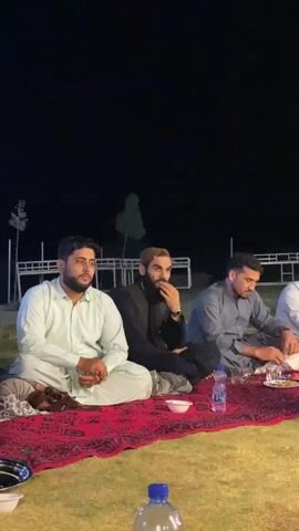 The video shows a group of men sitting on the ground at night, likely for a meal or gathering. They are seated on a large, red patterned rug spread out on a grassy area. Several men are visible, all dressed in traditional South Asian attire. The man on the far left wears a light green shalwar kameez. Next to him, a man with a beard and short blonde-tipped hair wears a black outer garment over a lighter shirt. To his right, a man in a blue shalwar kameez is seen interacting with food and drinks on a plate. Further to the right, another man in a white shirt is also present, his face partially obscured. A bottle of water and some dishes are placed on the rug in front of the men. In the background, there are fences and some sparsely planted trees, illuminated by overhead lighting, indicating an outdoor setting. The overall atmosphere appears to be relaxed and social.