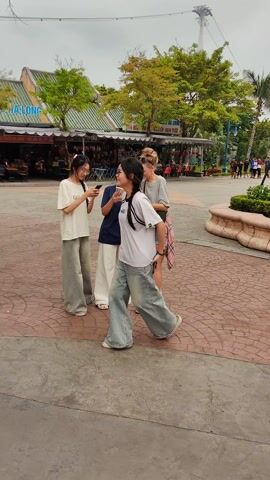 The scene unfolds in an outdoor, public space, likely a park or recreational area, paved with patterned brick or stone. Three young women stand in a small group, all focused on a smartphone held by one of them. A fourth woman walks away from the group.

The woman holding the phone is wearing a navy blue polo shirt and white trousers. The woman on her left has a white t-shirt and light-wash, wide-legged jeans, her long dark hair partially pulled back. The woman on the right is wearing a grey t-shirt, denim shorts, a pink plaid shirt tied around her waist, and sneakers. She also has a small crossbody bag. Her blonde hair is tied up in a bun.

In the background, there are commercial stalls or small shops, trees, palm trees, and a distant structure that appears to be a tower or ride. Other people can be seen walking in the background. There is a stone planter or fountain nearby. The sky appears overcast.