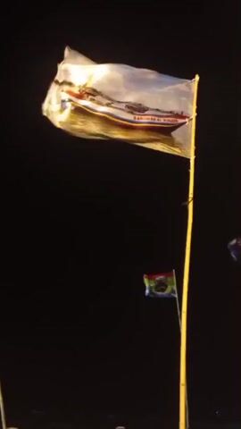A long, thin flag with an image of a boat is flying on a pole against a dark background. Below it, a smaller flag with a colorful emblem is also visible. The larger flag appears to be made of a translucent material and is illuminated, revealing the boat image and some text. The scene suggests an outdoor setting at night or in low light conditions.