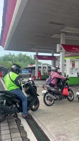 The video shows a busy scene at a Pertamina gas station (SPBU) during the daytime, likely in Indonesia. Several people on motorcycles are either waiting in line or actively refueling their vehicles.

In the foreground on the left, a person wearing a green jacket with a "Gojek" logo and a helmet is seated on a black scooter-style motorcycle, looking towards the gas pumps. This individual is positioned over a concrete drainage grate, with their left foot on the ground.

To the right, a line of motorcycles extends towards the gas pumps. A person wearing a pink long-sleeved top, a helmet, and carrying a purple backpack is on a silver underbone motorcycle, seemingly waiting their turn or preparing to refuel at pump 3. A red plastic bag hangs from the handlebars of their motorcycle. Behind them, another person in a pink t-shirt and shorts, without a helmet, is seated on a black scooter, also in line. Further to the right, another individual on a motorcycle is visible near pump 1.

The gas station features a large, grey canopy covering the pumps. Three main pump islands are visible, labeled with large red numbers "1", "3", and "5".
- Pump island 3 has a red sign above it that reads "PERTALITE". Below this, a white sign says "PERTALITE PERTAMAX".
- Pump island 5 also has a white sign displaying "PERTALITE PERTAMAX".
- Pump island 1 has a large red sign indicating "PERTALITE PERTAMAX TURBO" and a white sign with red text that says "JALUR KHUSUS MOTOR" (Special Lane for Motorcycles) with an arrow pointing down, suggesting a dedicated fueling spot for motorcycles.

All visible gas pumps are white with green and red accents, characteristic of Pertamina stations. A red fire extinguisher and a small blue plastic stool are placed near pump 1. In the background, a white building with a green roof is visible, and a banner with "IDUL FITRI" text can be seen on the left side of the background. The sky appears overcast. The ground is concrete, with a visible white circle marking on the ground near the black scooter.