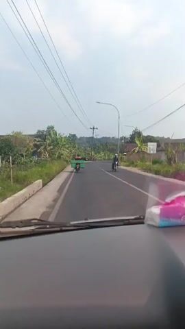 The video begins with a view from inside a vehicle looking out through the windshield onto a two-lane paved road. The sky is overcast, and there are power lines running parallel to the road. On the left side of the road, there is a grassy embankment with banana trees and some buildings. On the right side of the road, there are more trees and buildings. There are several motorcycles on the road, some with riders and some carrying cargo. One motorcycle in the distance on the left has a green cargo box. A black van passes on the right side of the road. The video appears to be taken while the vehicle is in motion, driving on the road.