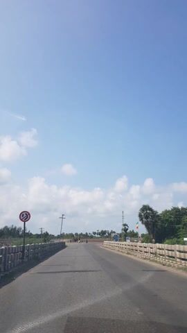 This media file is a video clip filmed from a moving vehicle on a sunny day in Zamin Budur, India. The scene is a rural highway with a clear blue sky and scattered white clouds. The road is paved and appears to be a two-lane highway with concrete barriers on either side. 

The video shows several vehicles, including cars and what appears to be a truck or construction vehicle. There are also utility poles with wires running along the side of the road, and trees, including palm trees, dot the landscape. In the distance, there seems to be some construction work in progress, with mounds of dirt and barriers visible.

A circular sign with red lettering and a white background is visible on the left side of the road. The sign appears to be a traffic warning or regulatory sign. The overall mood of the video is calm and captures a typical day's journey through a rural Indian landscape. The time of day appears to be late morning or early afternoon, given the bright sunlight and the length of the shadows. The weather is fair and pleasant.