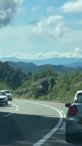 The video shows a bright daytime scene on a winding mountain road. In the foreground, the rear of a silver Volkswagen Polo is visible, with something dark (possibly a piece of clothing or a bag) hanging out of its trunk. The car's license plate shows "RADOMAN AUTO" in red. Ahead of it, a white Volvo SUV is driving around a bend, and another white car is further in the distance. The road has double white lines, indicating no overtaking. On the left side of the road, a circular red sign with a white bar, possibly a "no entry" or "do not enter" sign, is visible. The road is surrounded by steep, lush green mountains covered in trees and some rocky outcrops. The sky is partly cloudy with patches of clear blue. The overall impression is a scenic drive through a mountainous region. Background music is playing throughout the video. The location is contextualized as Dobrsko selo, Montenegro.
