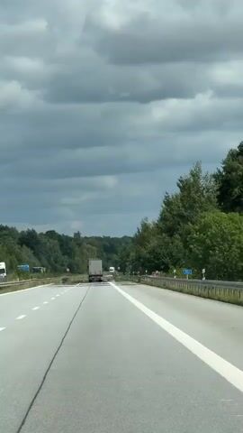 A dashcam perspective from a vehicle driving on a highway. The road is paved with asphalt and has white lane markings. On the right side of the road, a metal guardrail is visible, and beyond that, a dense forest line. On the left side of the road, trees and vegetation are also visible, with some areas appearing overgrown. There are traffic signs and posts along the roadside. In the distance, a white semi-truck is traveling in the same direction as the camera vehicle. The sky above is cloudy with a mix of gray and white clouds. The overall environment suggests a rural or semi-rural highway setting on a possibly overcast day.